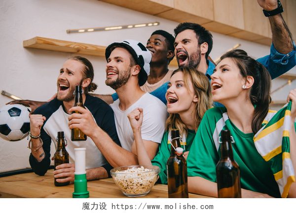 一群年青人喝着啤酒在观看足球比赛兴奋多文化的朋友与啤酒瓶做是手势在观看足球比赛在酒吧 